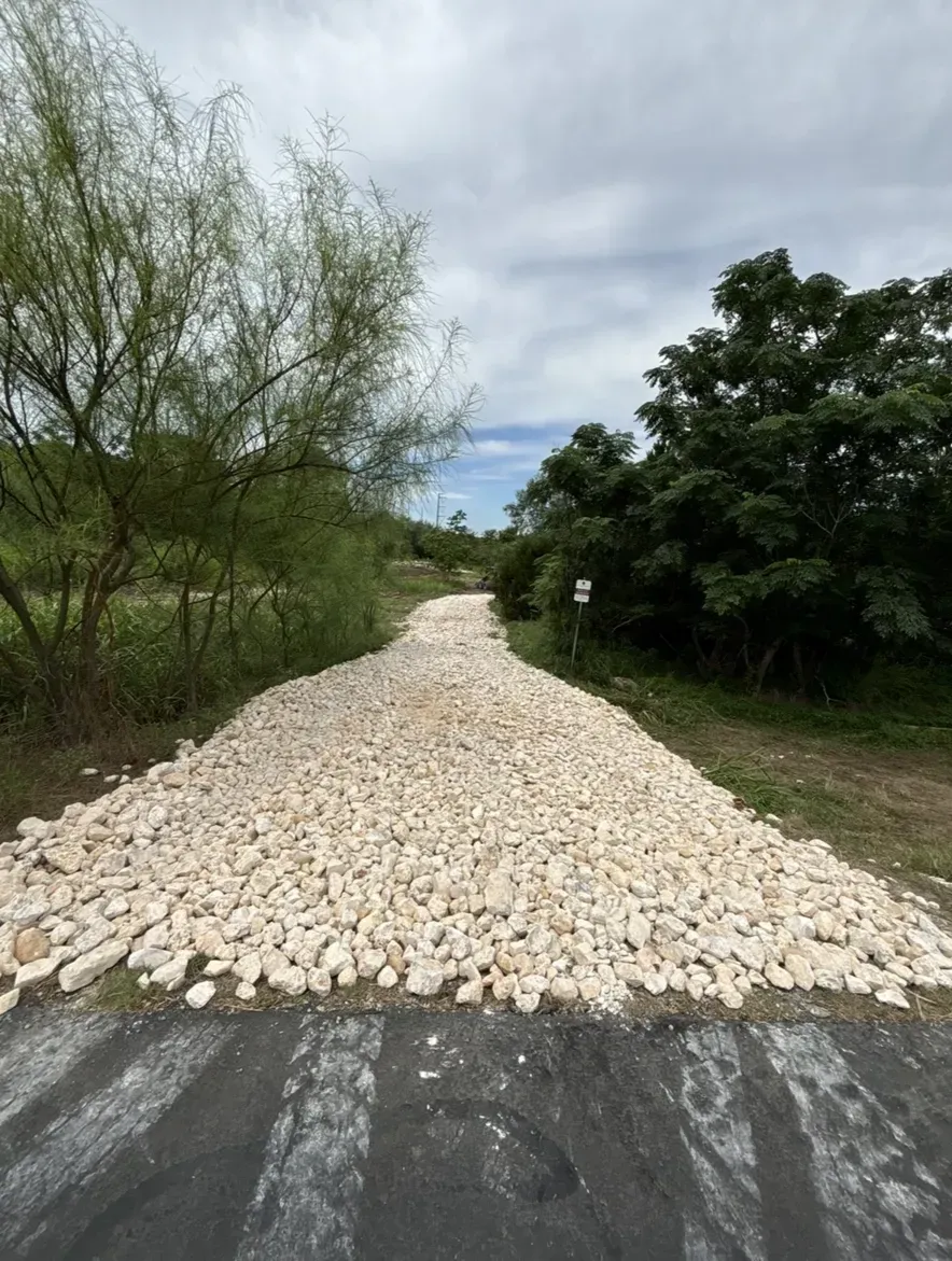 A gravel-covered path on a paved road, bordered by green trees under a cloudy sky.