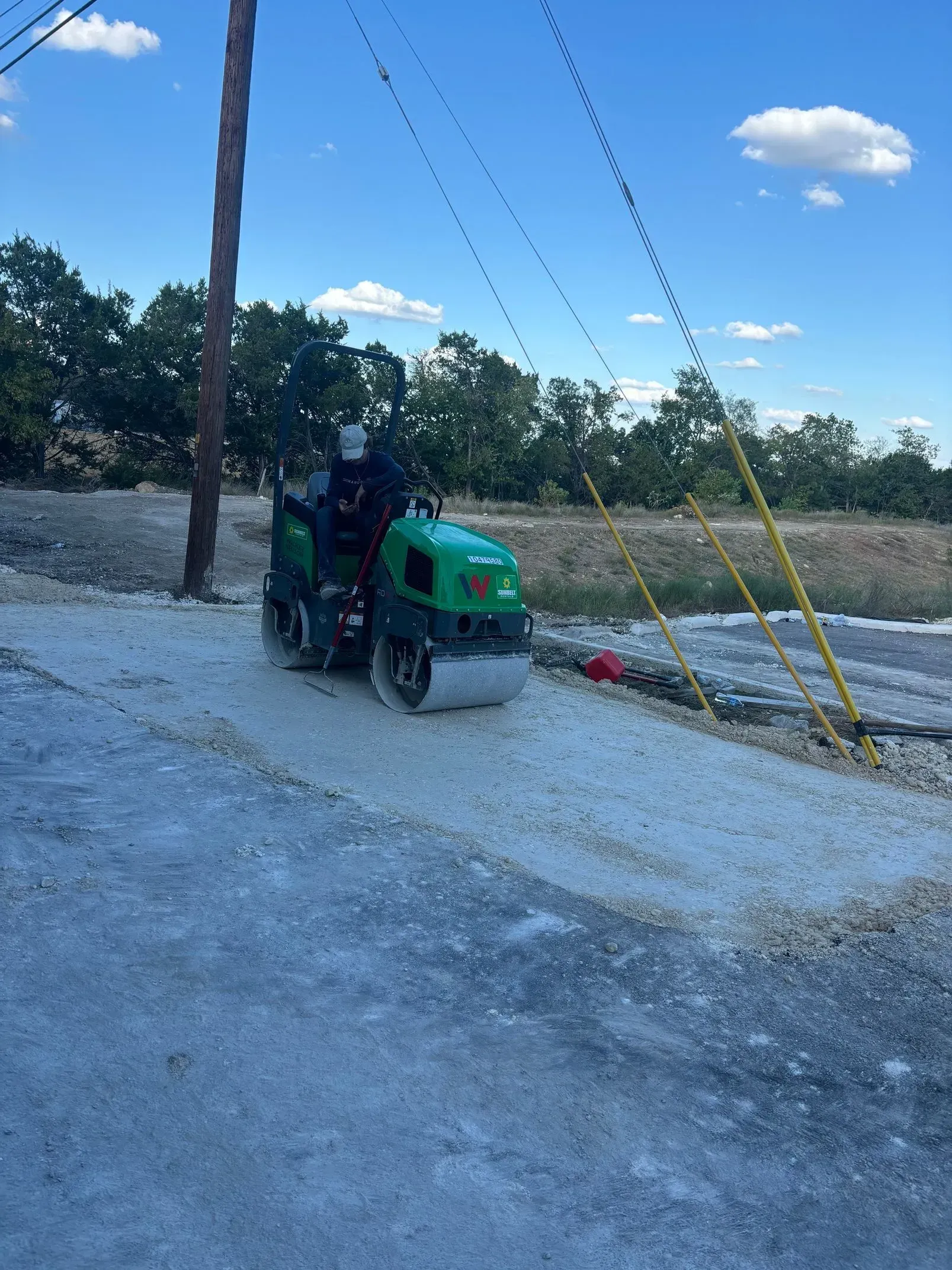Green roller compacting gravel road, operator in cab, power lines overhead.