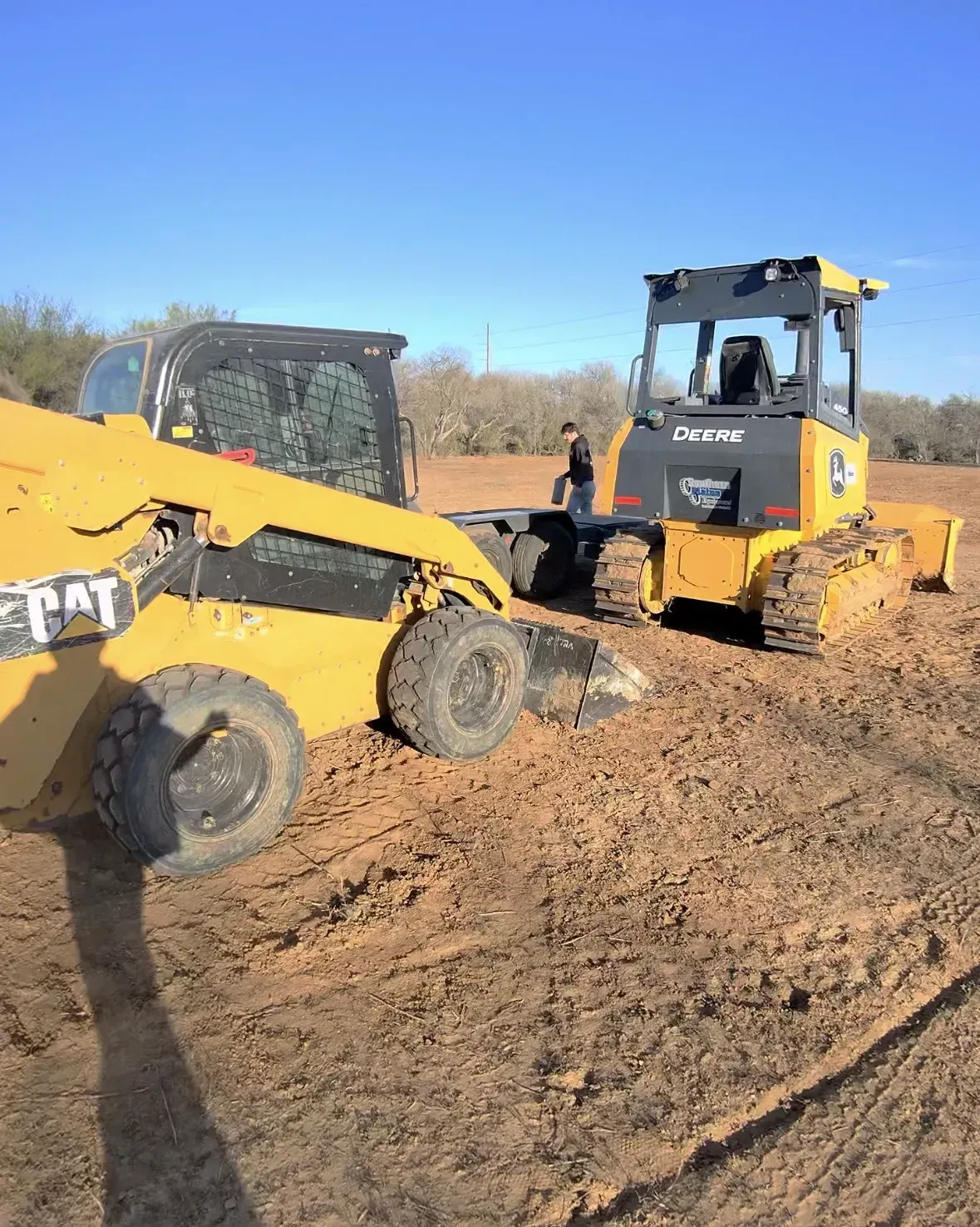 Yellow CAT skid steer pulling a yellow bulldozer on a dirt field under a blue sky.