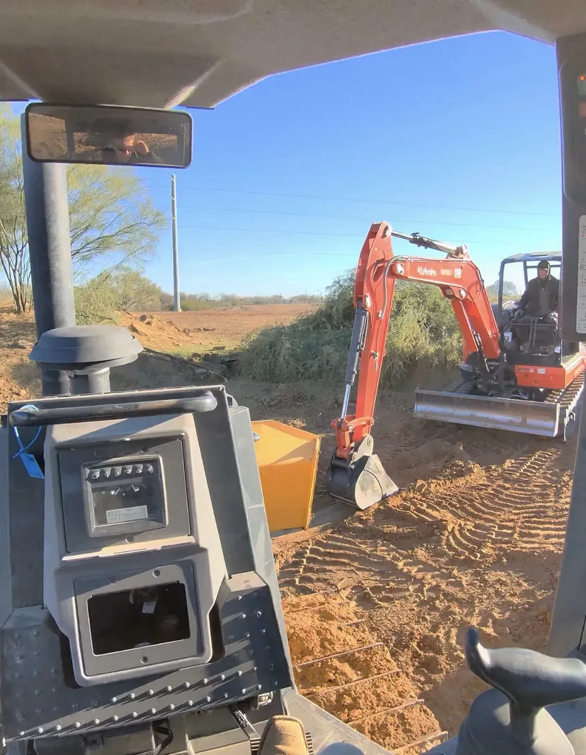 View from inside a machine with a small orange excavator digging dirt under a clear sky.