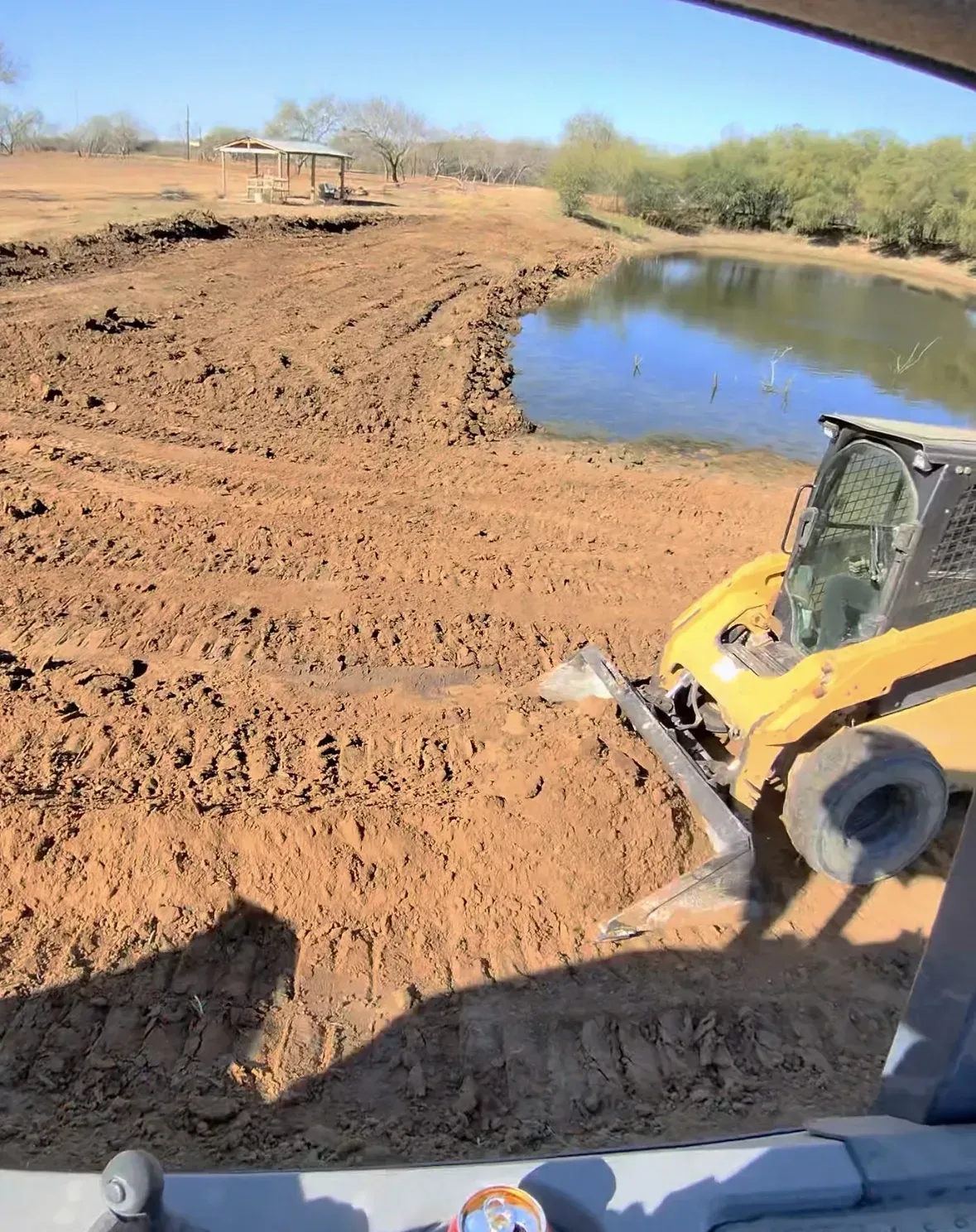 Yellow skid steer working on muddy land next to a pond.