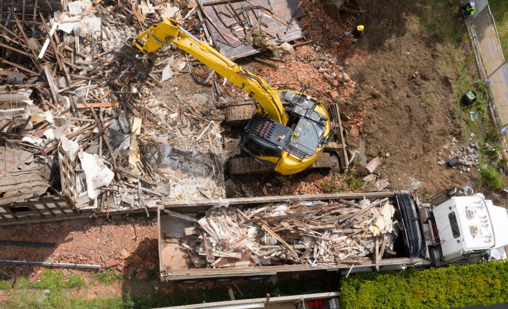 A yellow excavator demolishes a multi-story brick building on a sunny day.