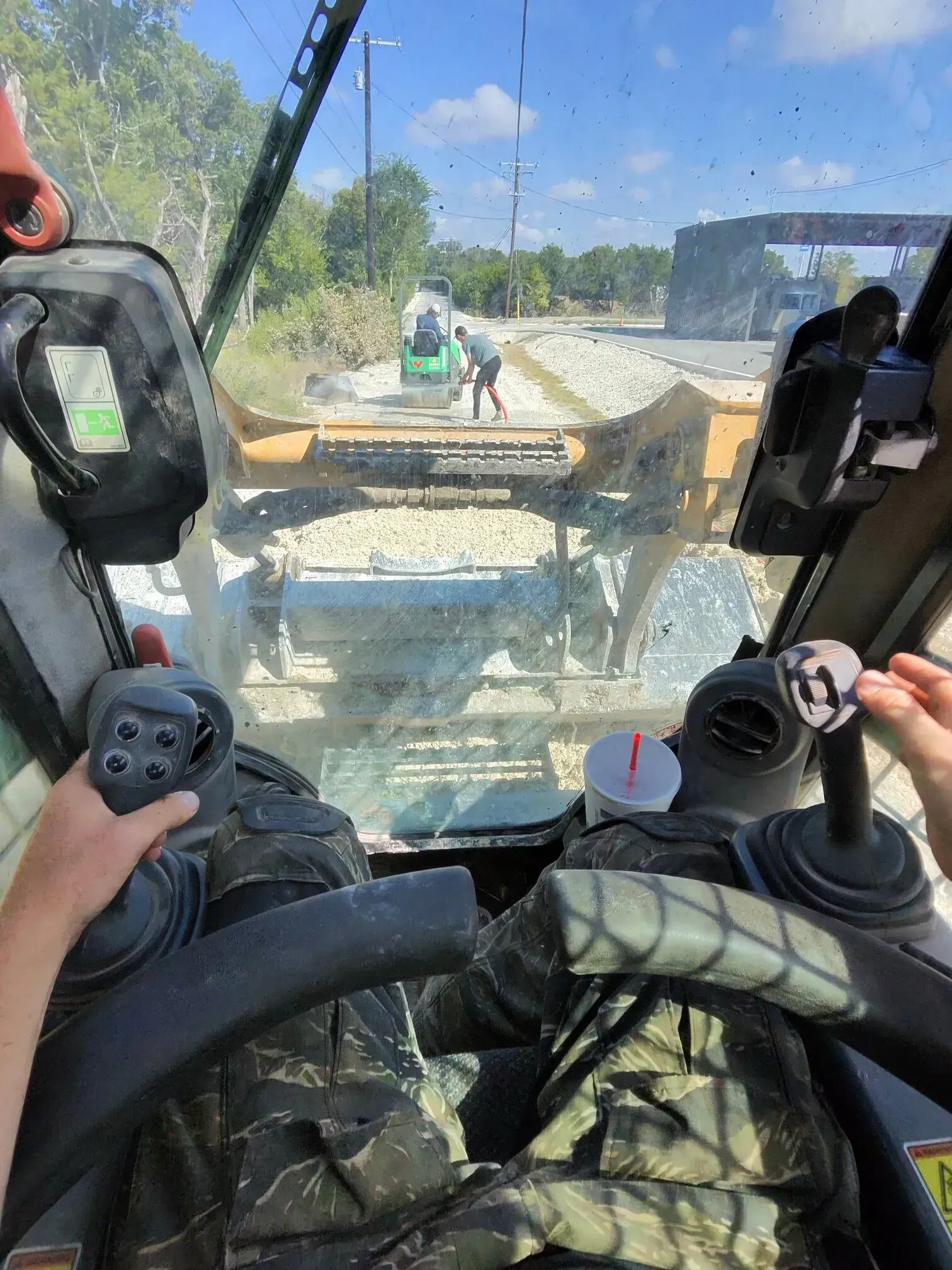 View from inside a skid steer operating on a road construction site; person operating controls.