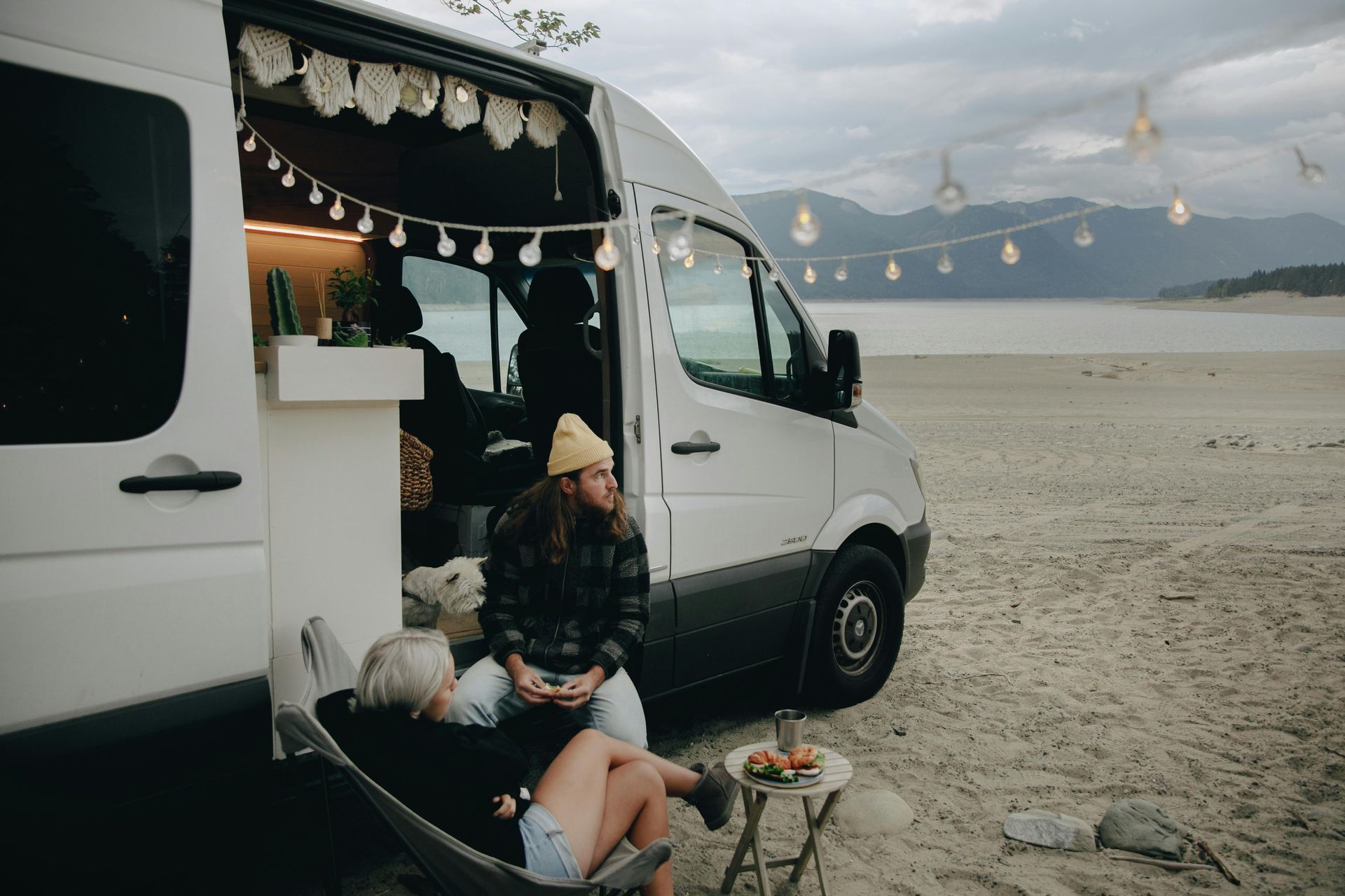 Two people relax by a van on a beach, lit by string lights. Mountains in the background.