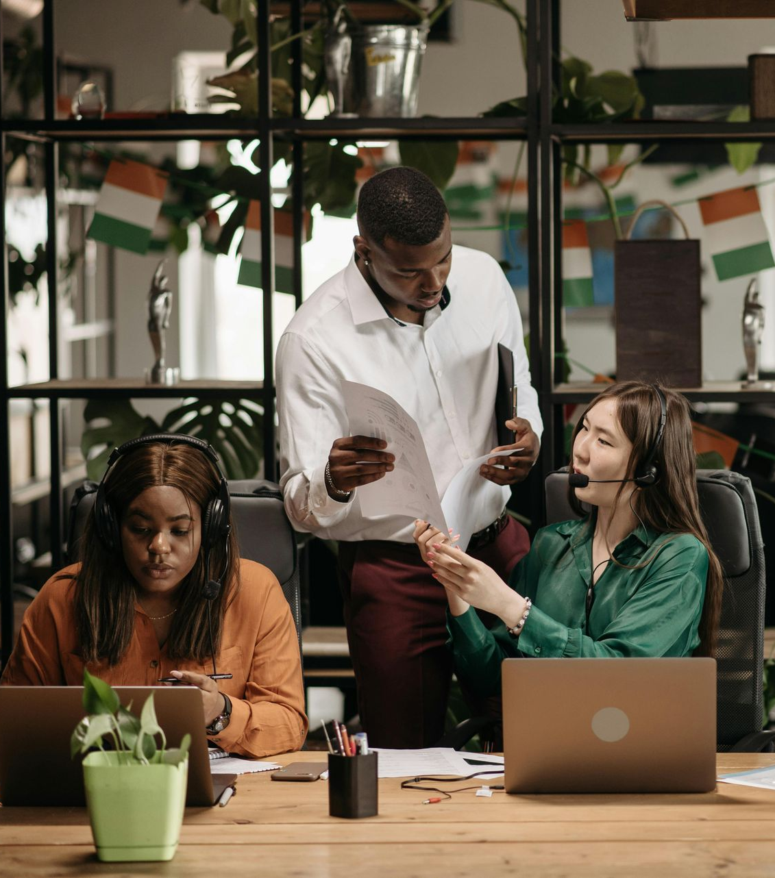 Three office workers collaborate around a desk with laptops and paperwork in a workspace decorated with green plants.