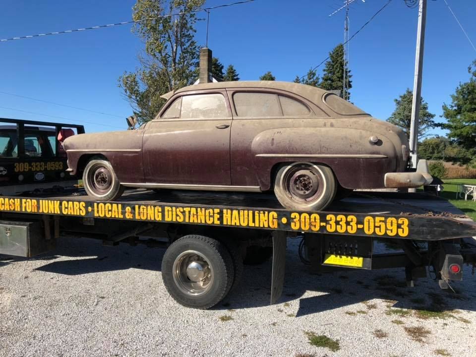 Dusty maroon classic car on a flatbed tow truck. The car is outdoors on a sunny day.