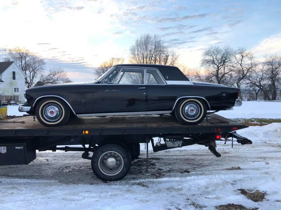 Black classic car on a tow truck, parked in a snowy rural area.