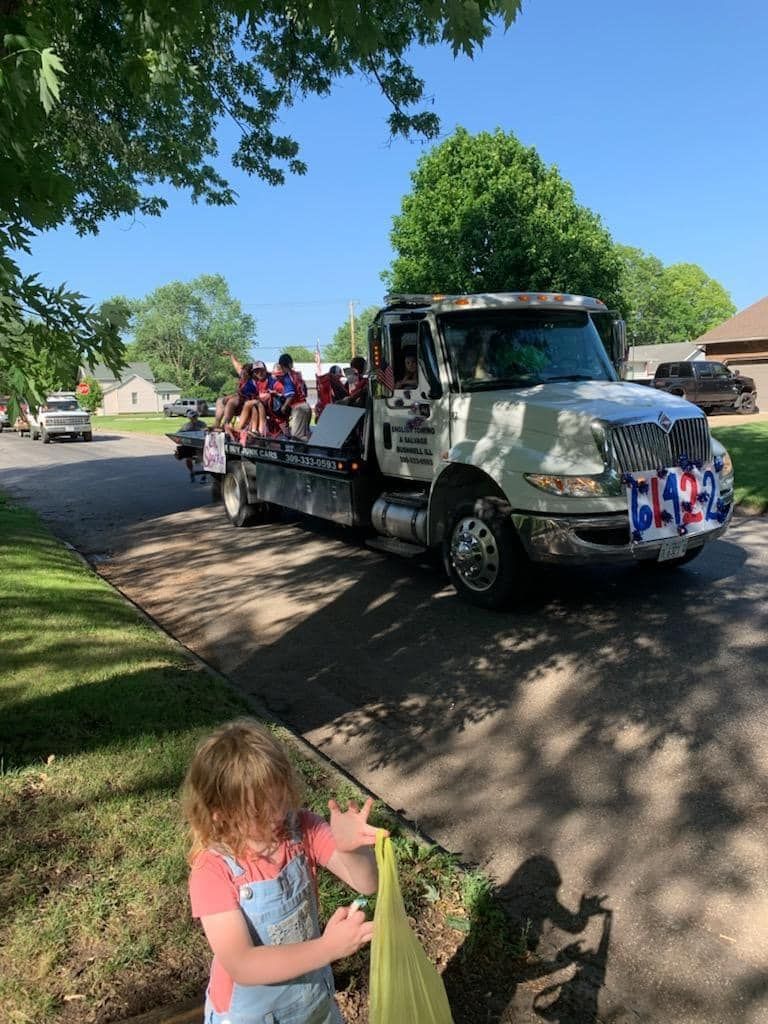 A child watches a parade from a residential yard. A flatbed truck carries people, decorated with red, white, and blue.