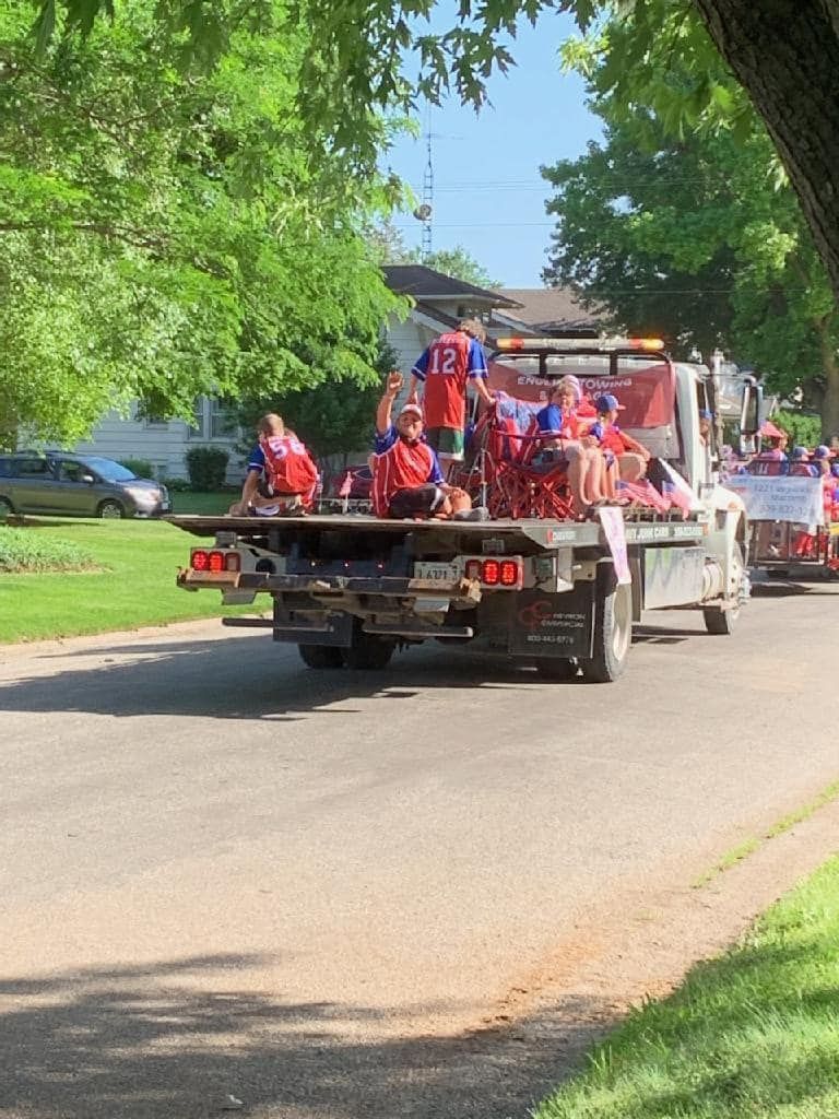 People on a truck bed float in a parade, red and blue attire, sunny day.