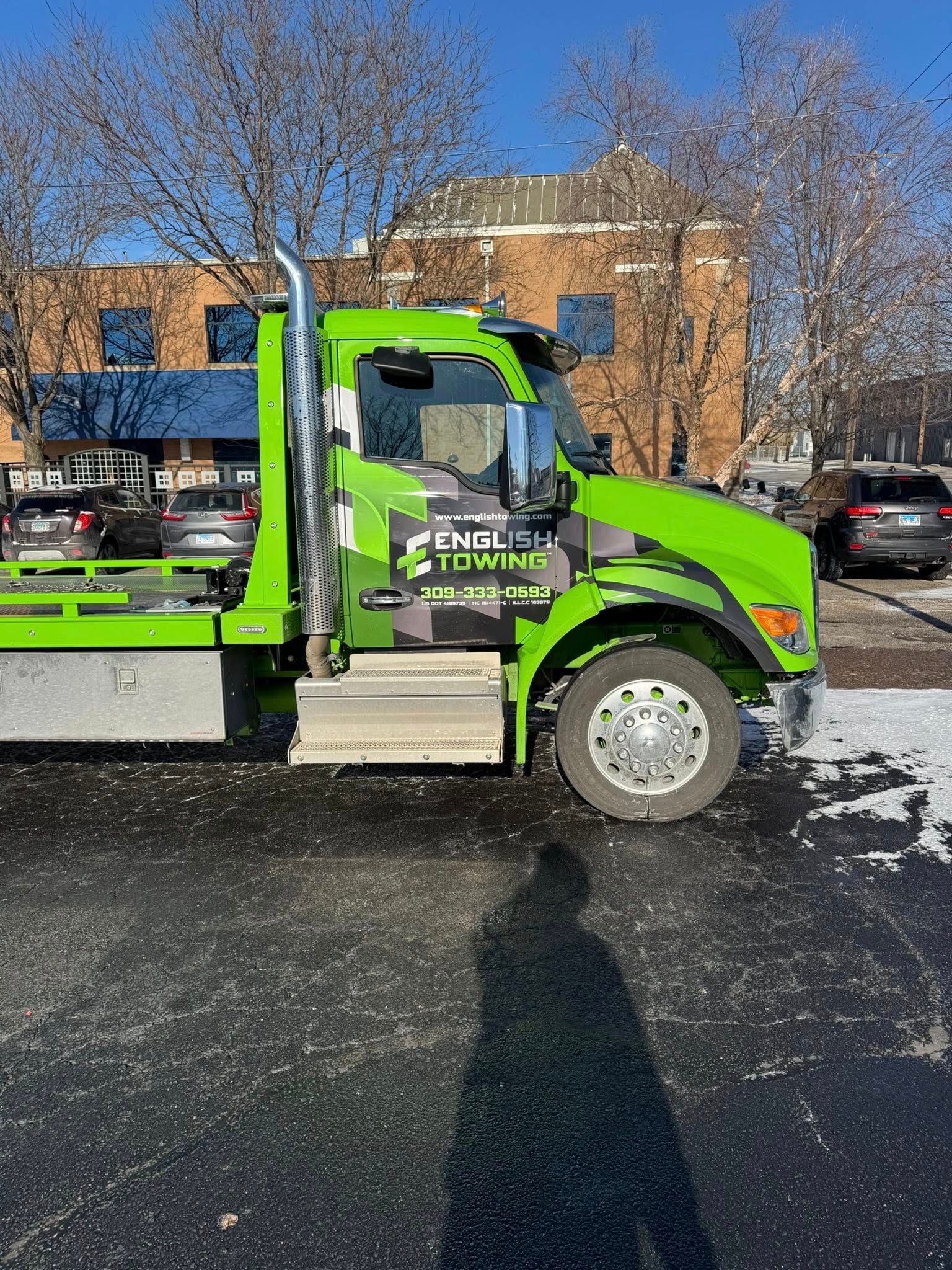 Green tow truck with business logo parked on asphalt. Sunny day with building in background.