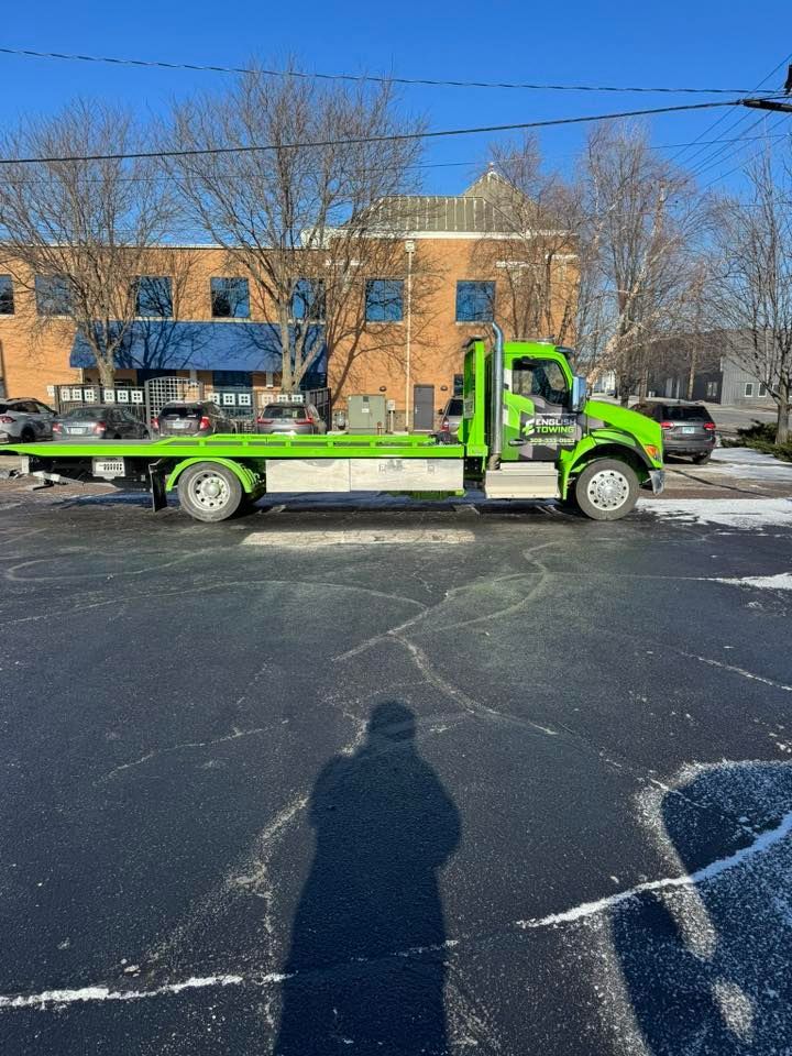 Green flatbed tow truck parked on asphalt. Shadow of a person is in the foreground. Building in the background.