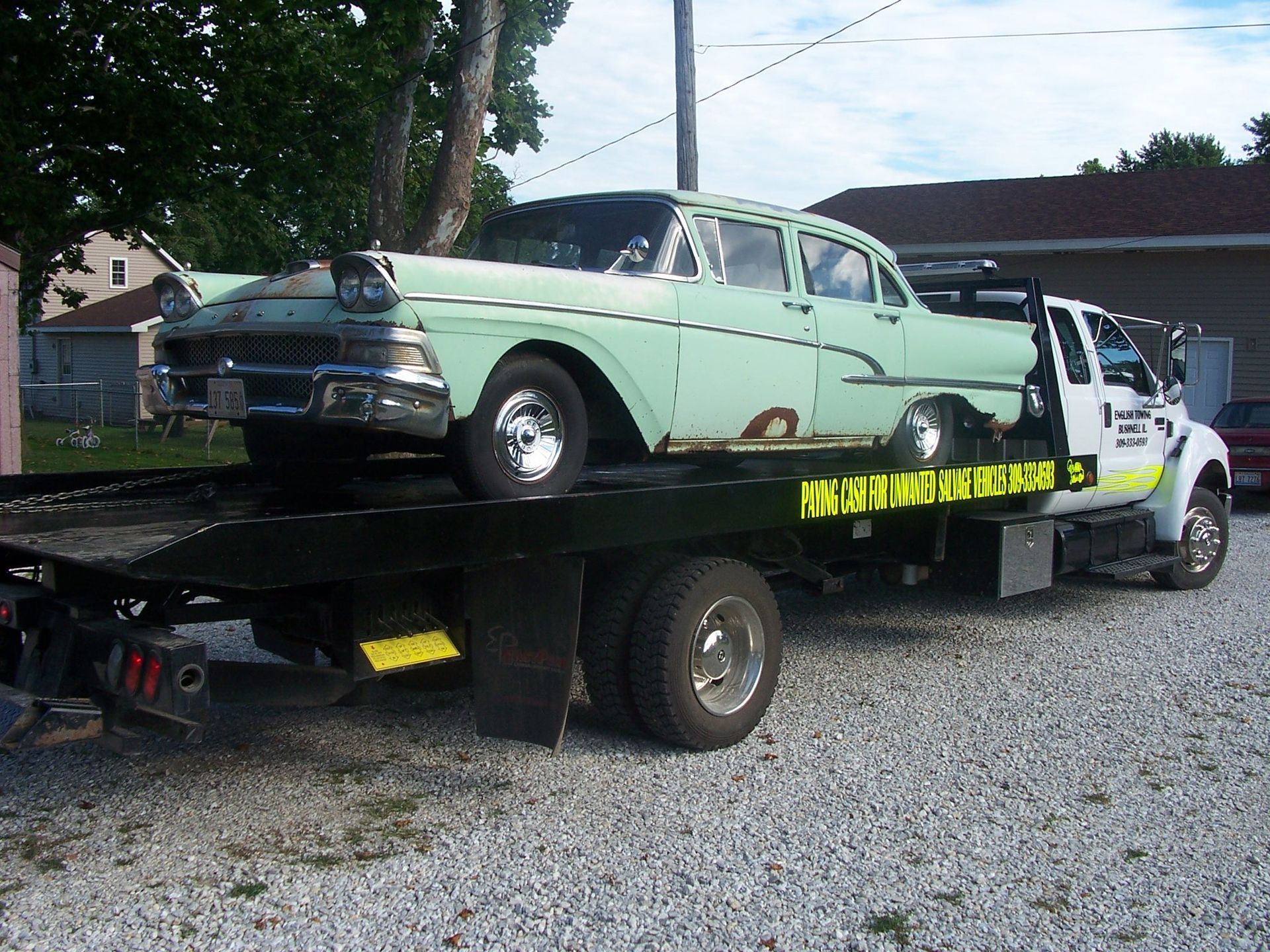 A vintage green car is loaded onto a tow truck on a gravel driveway.