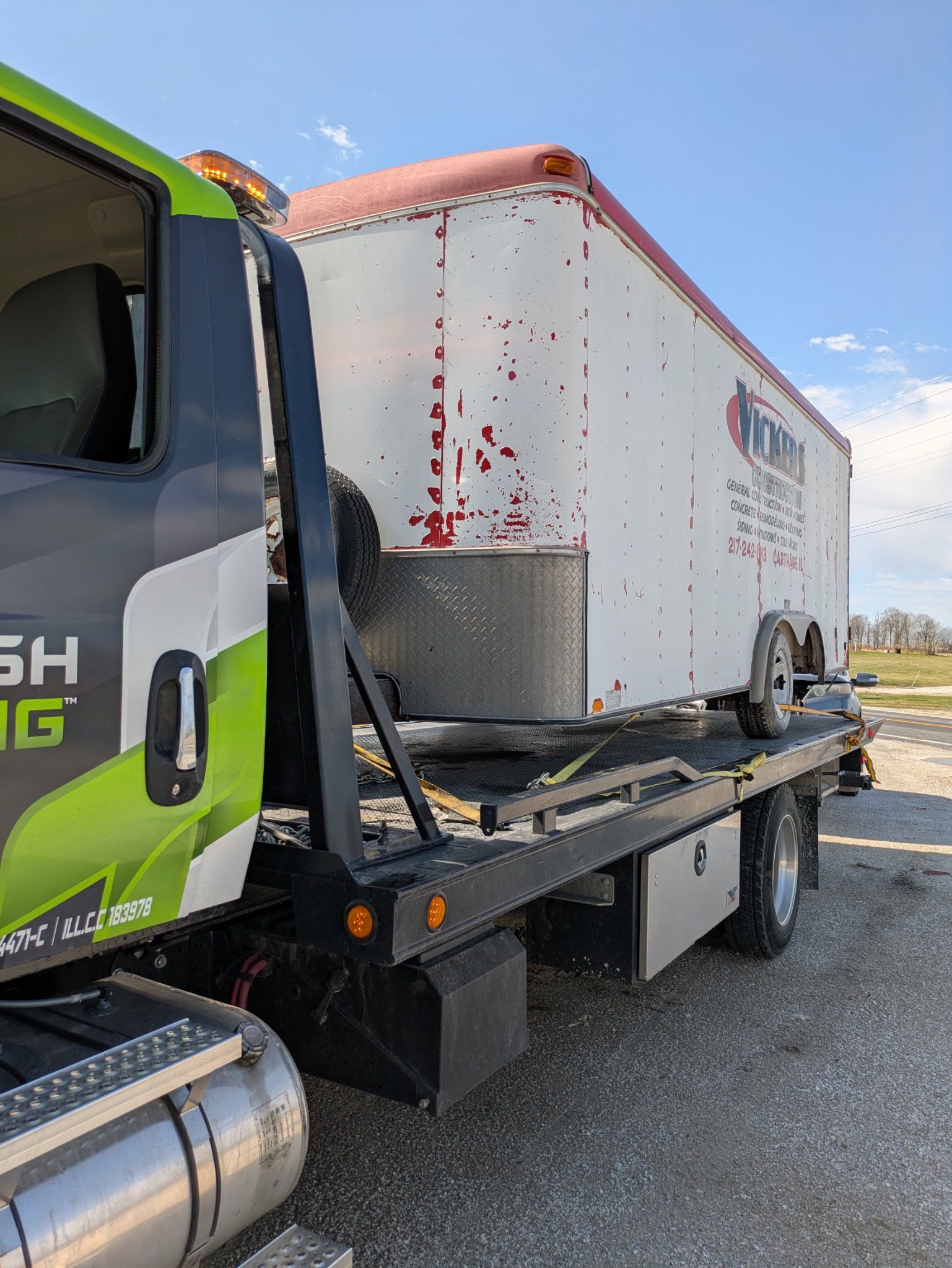 Tow truck hauling a white box trailer with red trim on a sunny day. The truck has green and black decals.
