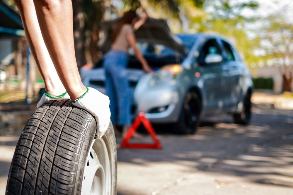 Person changing a car tire on the side of a road, another person with the hood of the car open.
