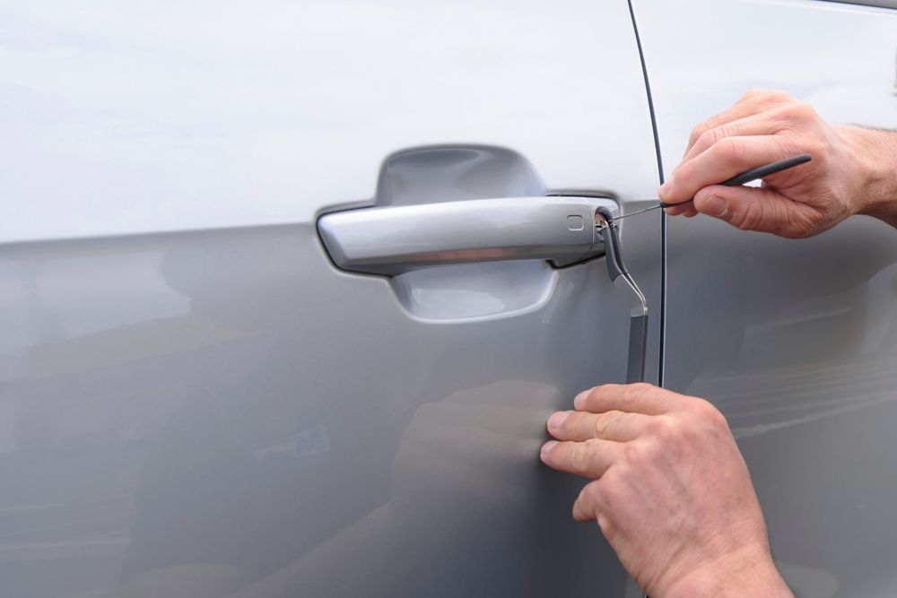 Hands using lock picking tools on a silver car door handle to unlock it.