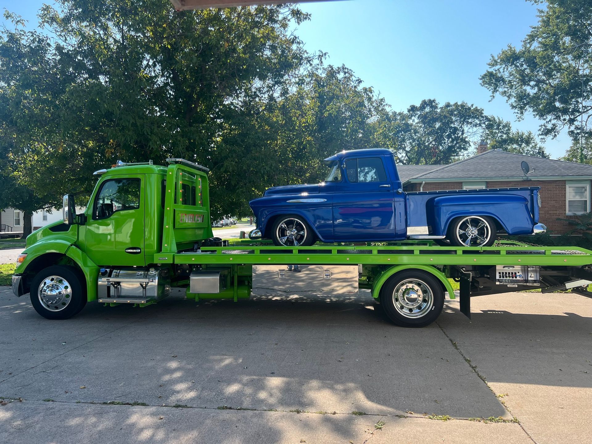 Green tow truck carrying a blue classic pickup truck on a sunny day.