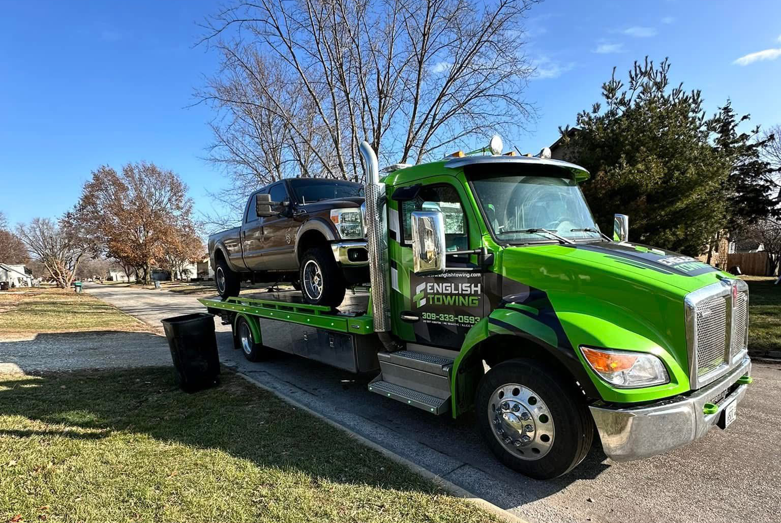 Green and silver tow truck hauling a brown pickup truck on a sunny day.
