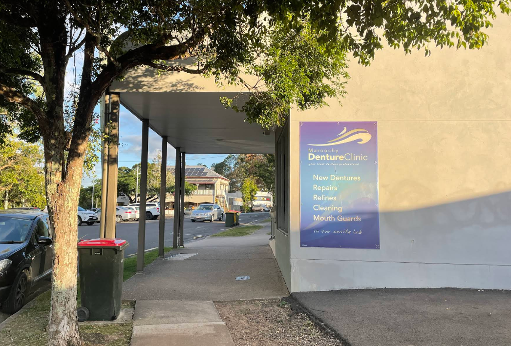 Sidewalk under a covered walkway, a sign for a business, and a street with parked cars. — Maroochy Denture Clinic In Yandina, QLD