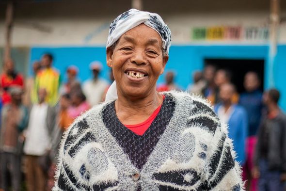 Woman smiles widely, wearing a patterned sweater and headscarf, in front of a blue building, other people blurred in background.