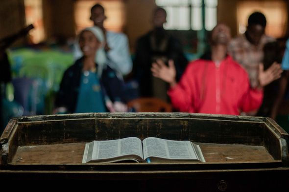 Open Bible on a wooden lectern, people in background raising hands, indoors.