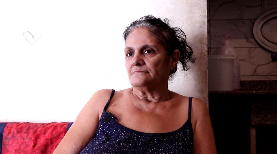 Woman in blue tank top looks at the camera. She sits against a white wall and red cushion.