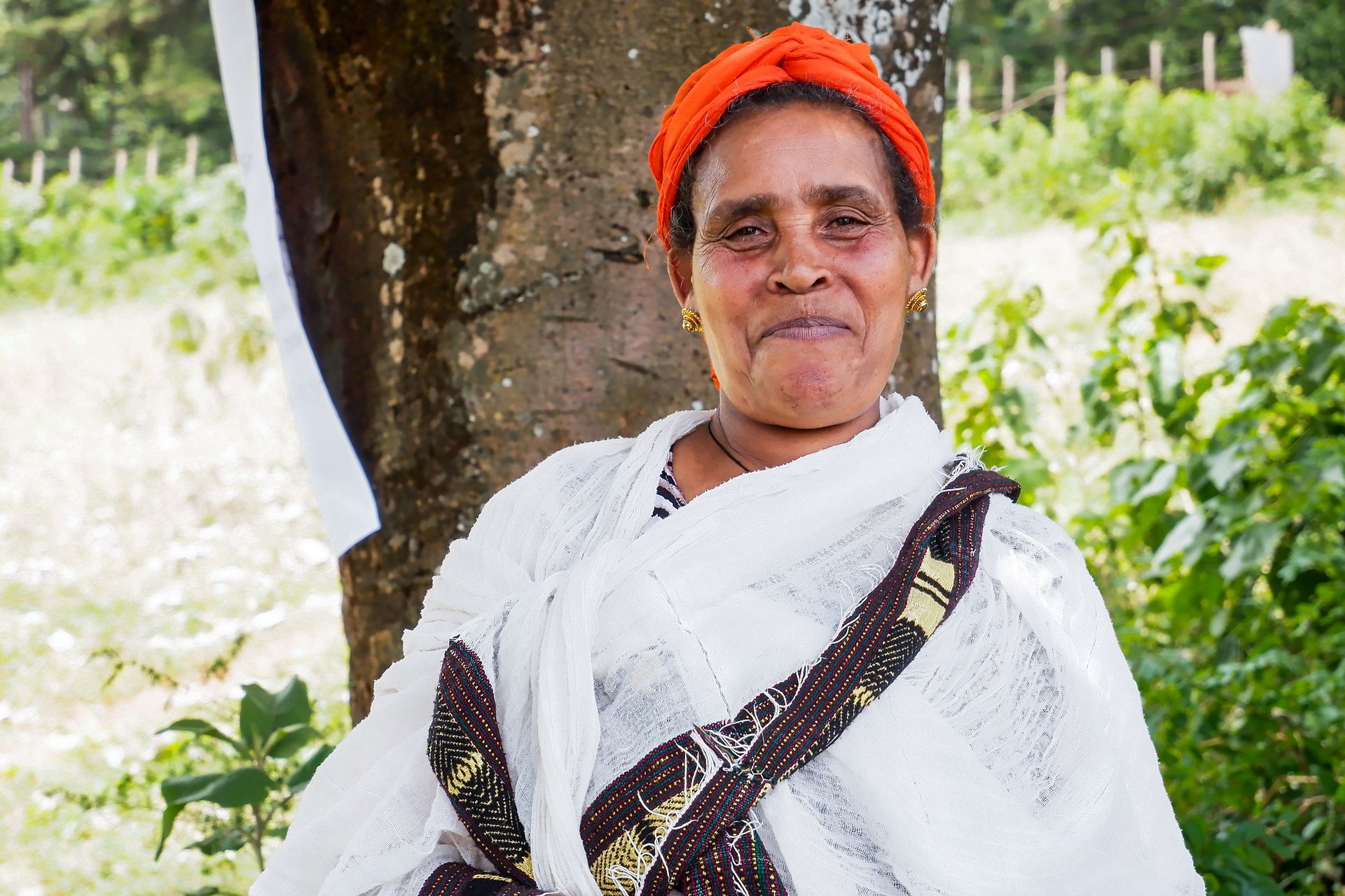 Woman in white with an orange headwrap smiles. Leans against a tree, with greenery in the background.