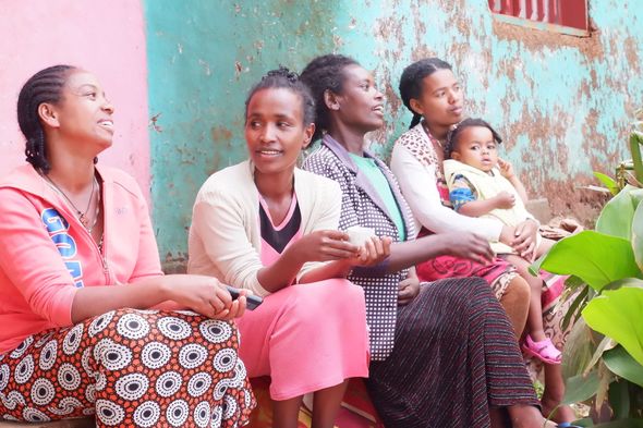 Five women and a child seated outside, talking, near a colorful wall and greenery.