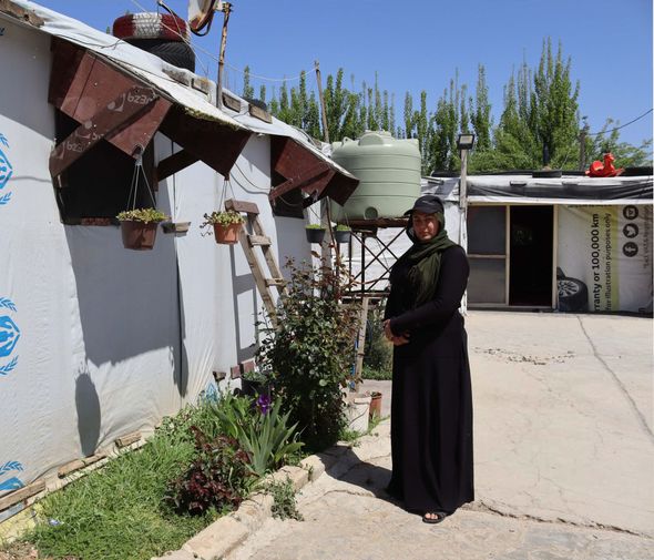 Woman in black stands in front of a modest building with a water tank and garden.