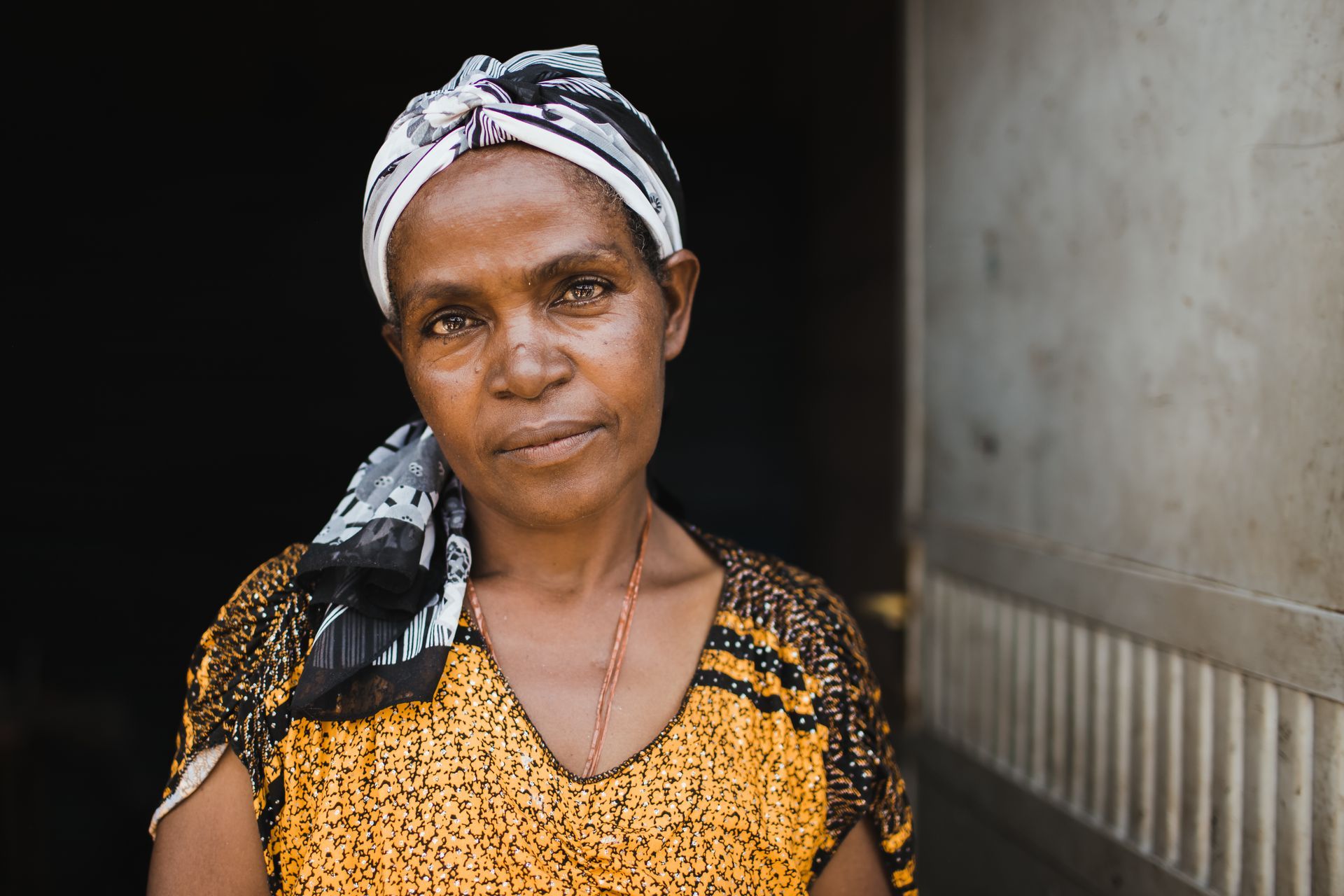 Woman wearing a patterned headscarf and yellow top, looking directly at the camera, standing in a doorway.