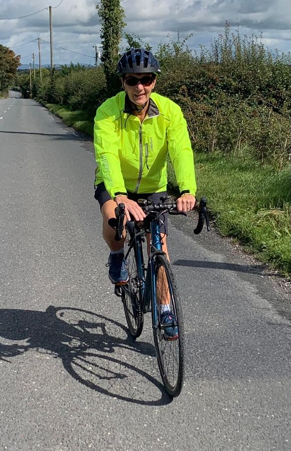 Cyclist riding on a paved road, wearing a bright yellow jacket and sunglasses, with a green landscape in the background.