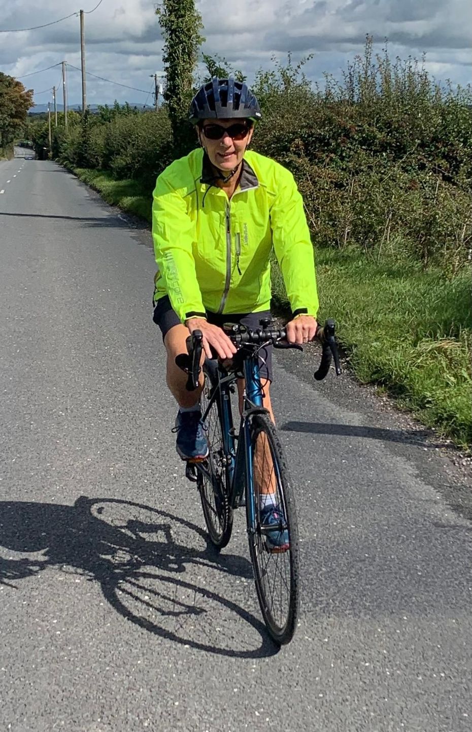 Cyclist riding on a paved road, wearing a bright yellow jacket and sunglasses, with a green landscape in the background.