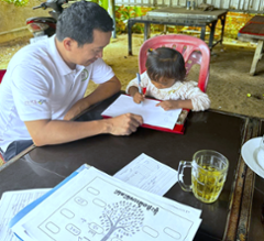 Man assisting a child with writing at a table, both focused on paperwork. Outside setting, documents visible.