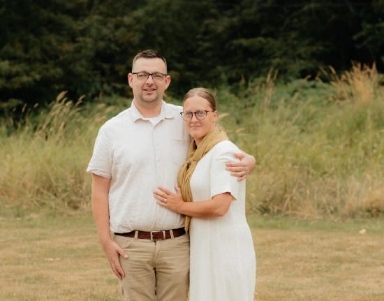 A couple stands outdoors, embracing. The man wears glasses, a light shirt, and khaki pants. The woman wears glasses and a white dress.