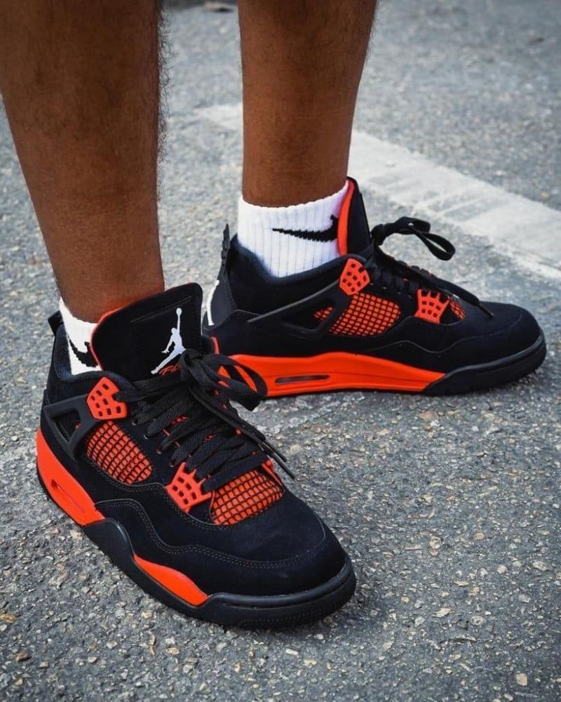 Black and orange Air Jordan 4 sneakers on a person's feet, white Nike socks, standing on pavement.
