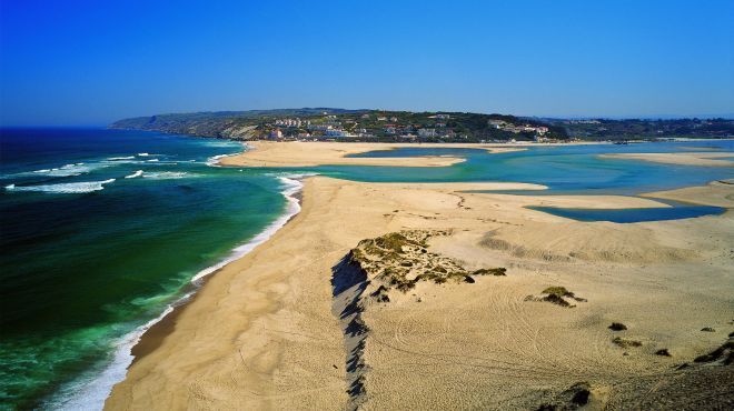 Vista aérea de uma praia de areia com água turquesa, ondas e uma formação rochosa sob um céu azul claro.