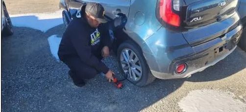 Person kneeling beside a gray car, tightening a wheel lug with a red tool on a gravel road.