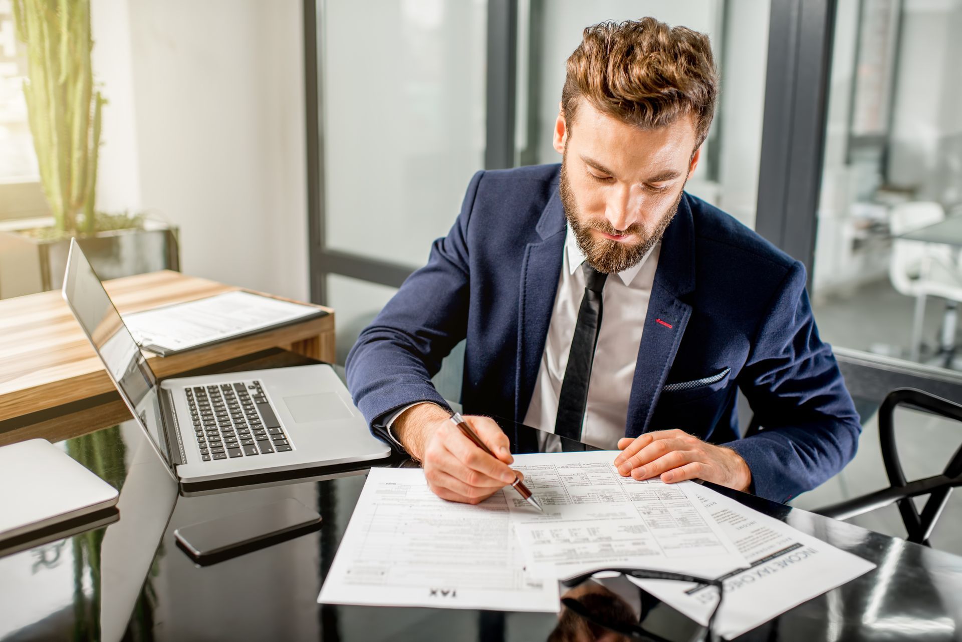 A tax accountant dressed in a suit, working with documents and a laptop in a modern office.