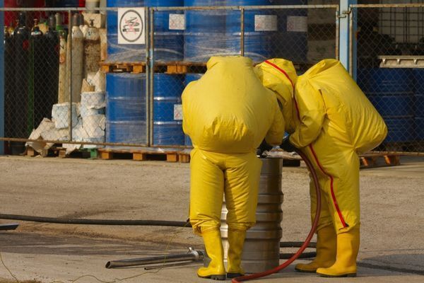 Two people in yellow protective suits are standing next to a barrel.