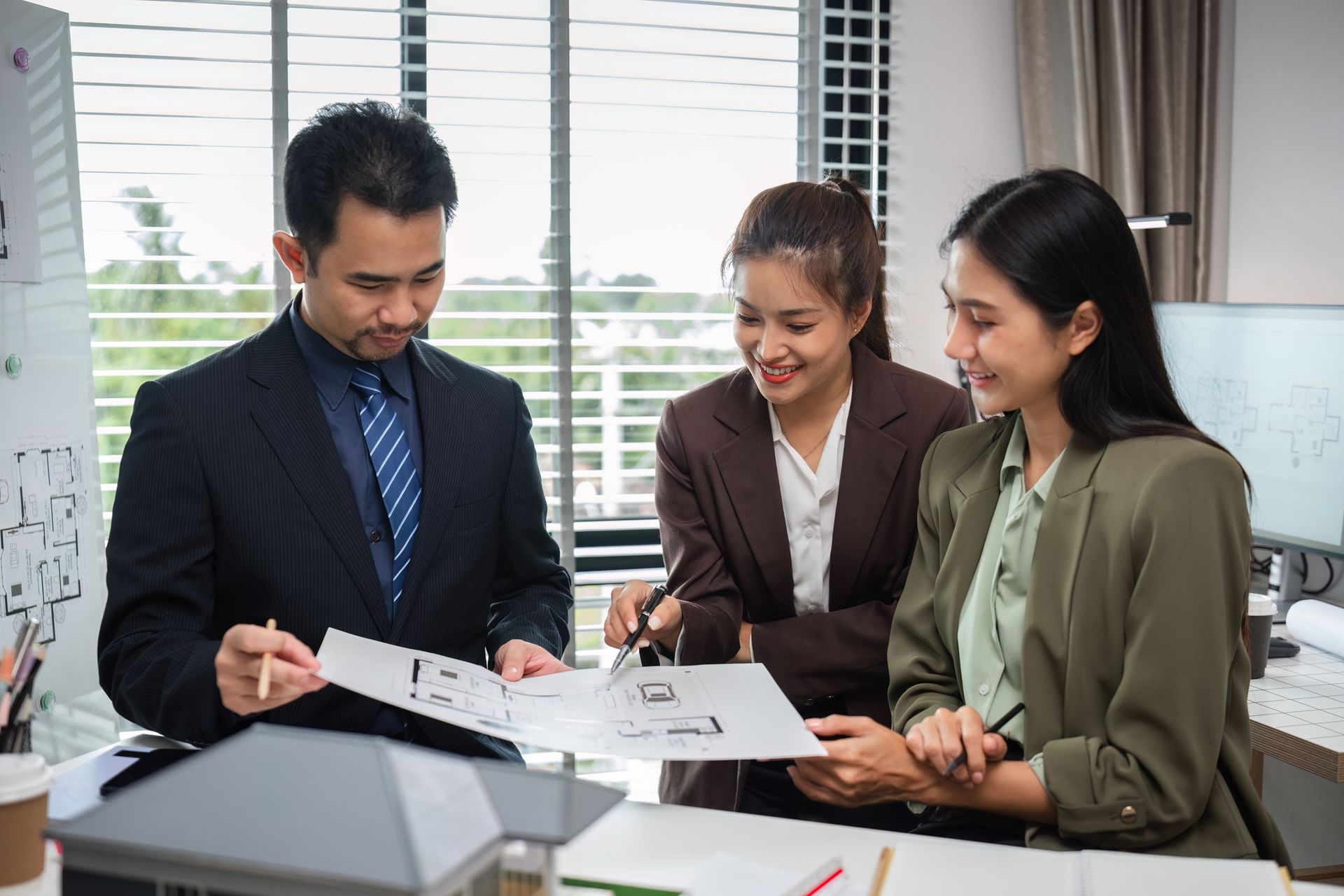 Three coworkers reviewing documents together at a desk in an office, with papers, laptop, and window blinds behind them