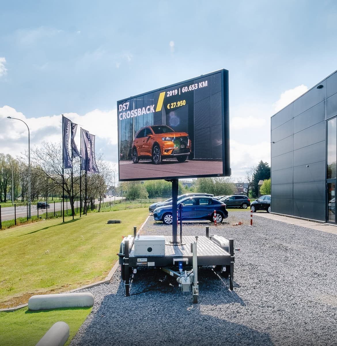 Een mobiel reclamebord toont een oranje SUV voor een autodealer. Blauwe auto's en een grindpad omringen het scherm met de aanhanger op een zonnige dag.