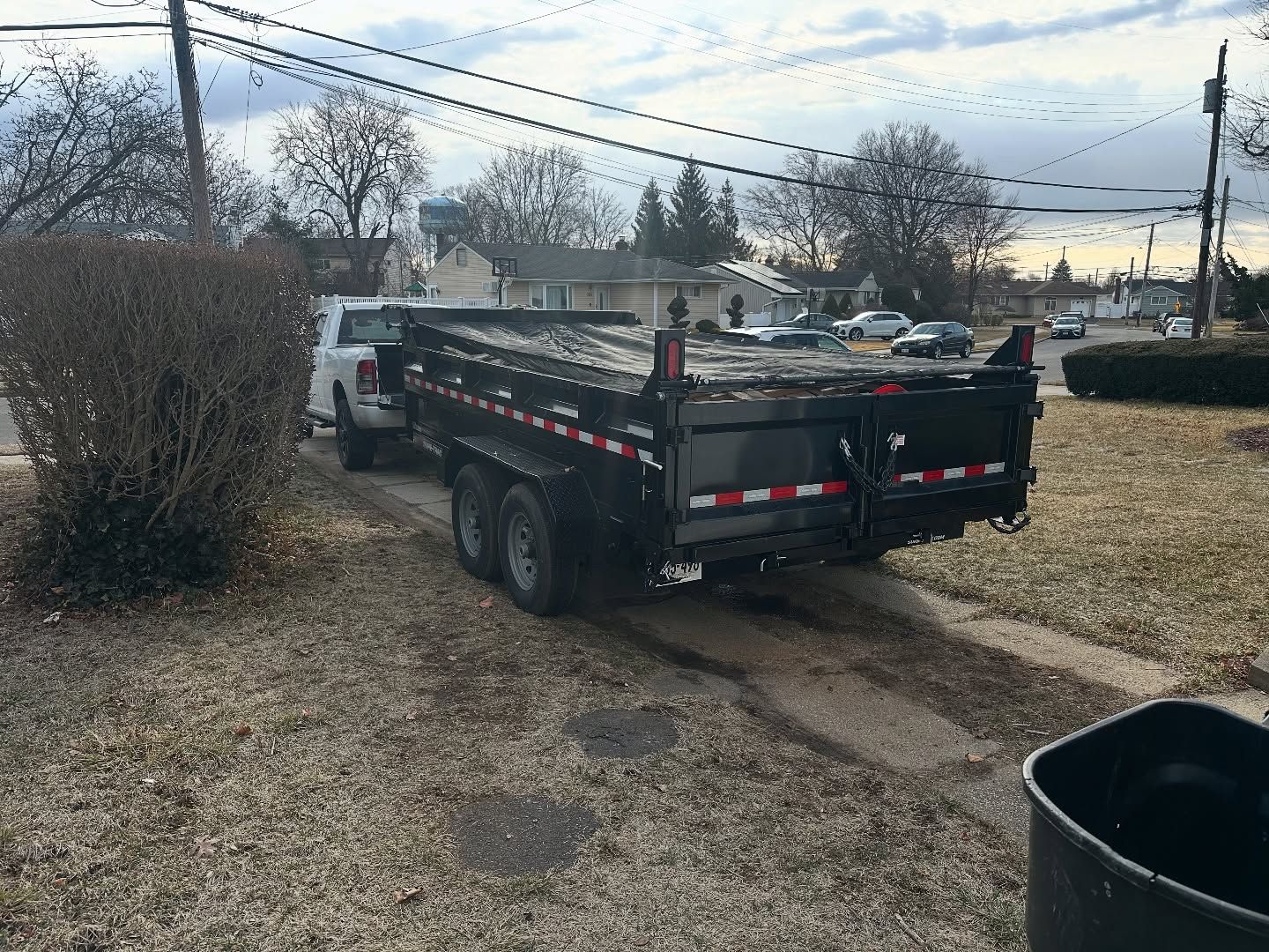 A white pickup truck pulling a black dump trailer on a driveway near a residential street.