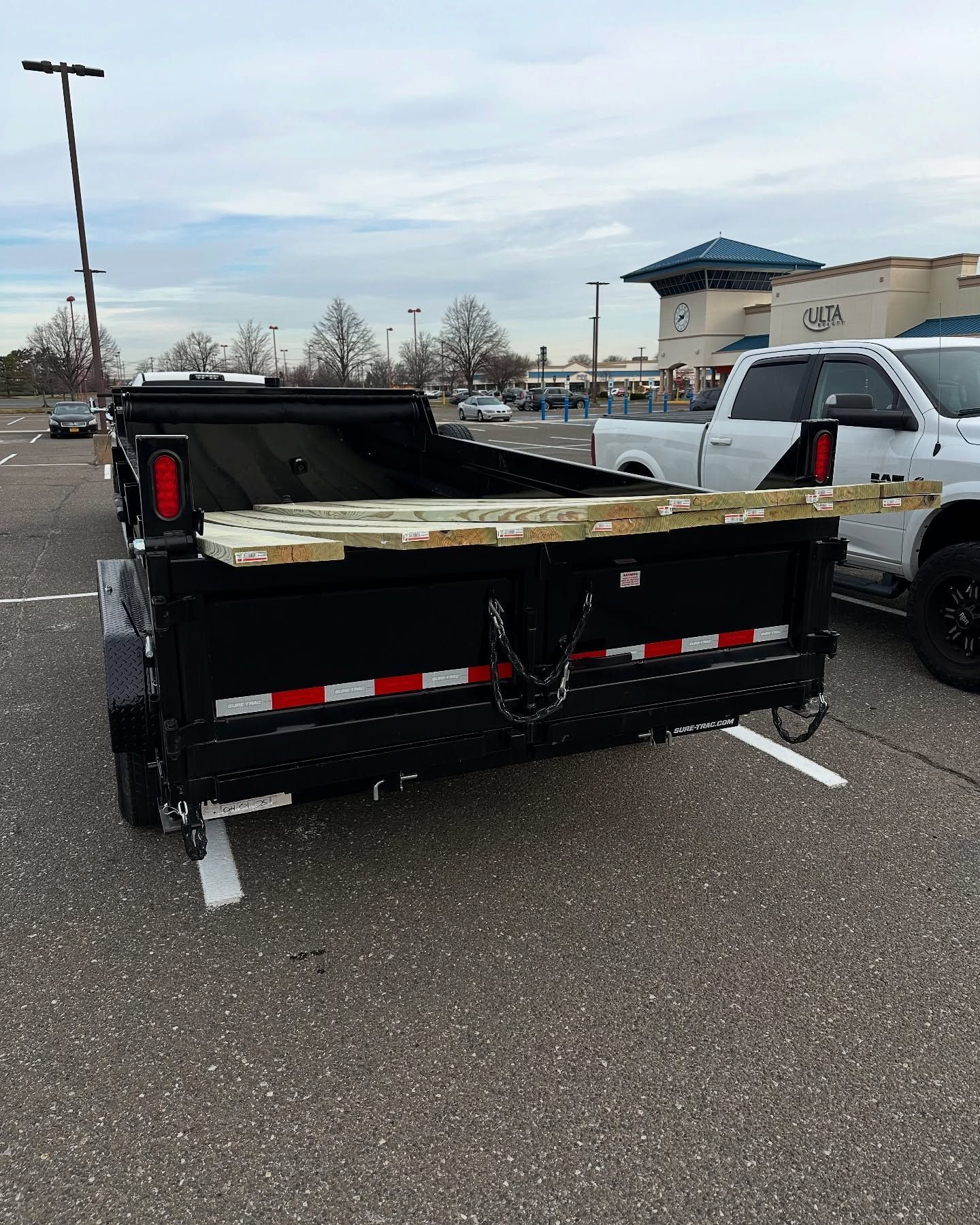Black trailer parked in a parking lot with wooden planks across the top. White pickup truck beside it.