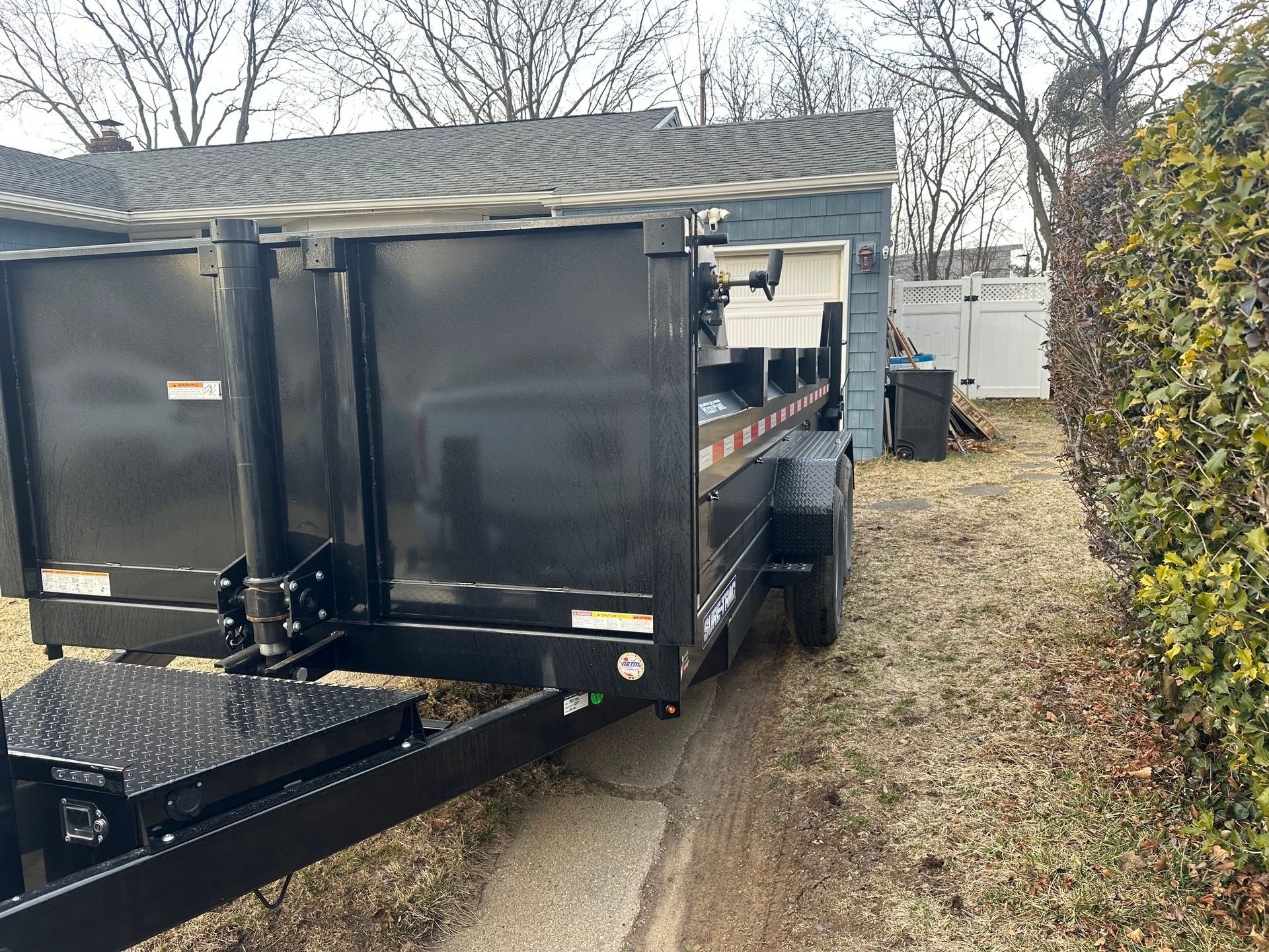 Black dump trailer parked on a patch of dirt next to a grassy area, with a house and bushes in the background.