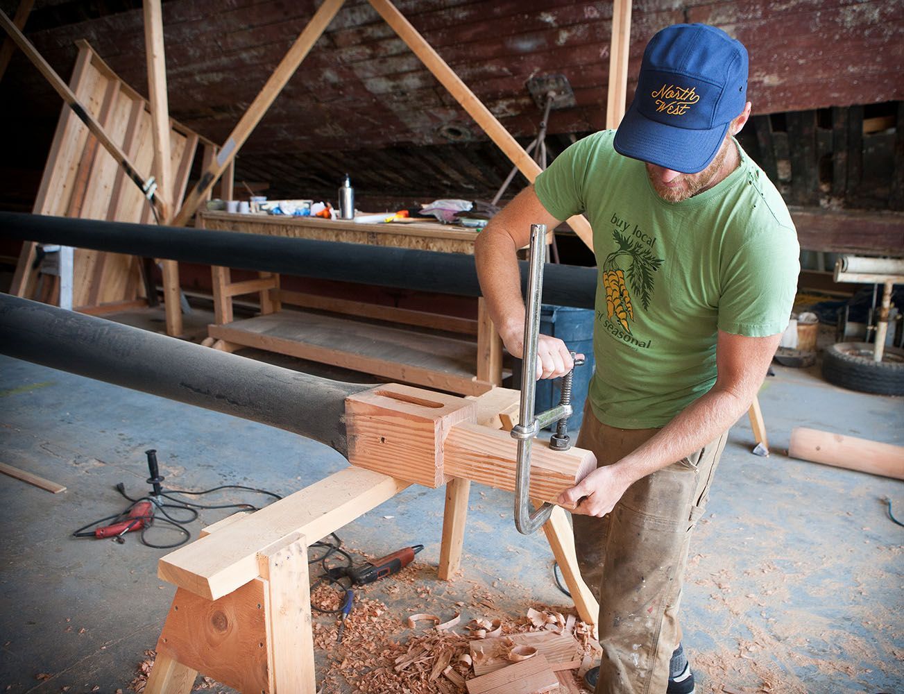 Man in blue cap uses clamp on wood piece attached to black pole in a workshop.