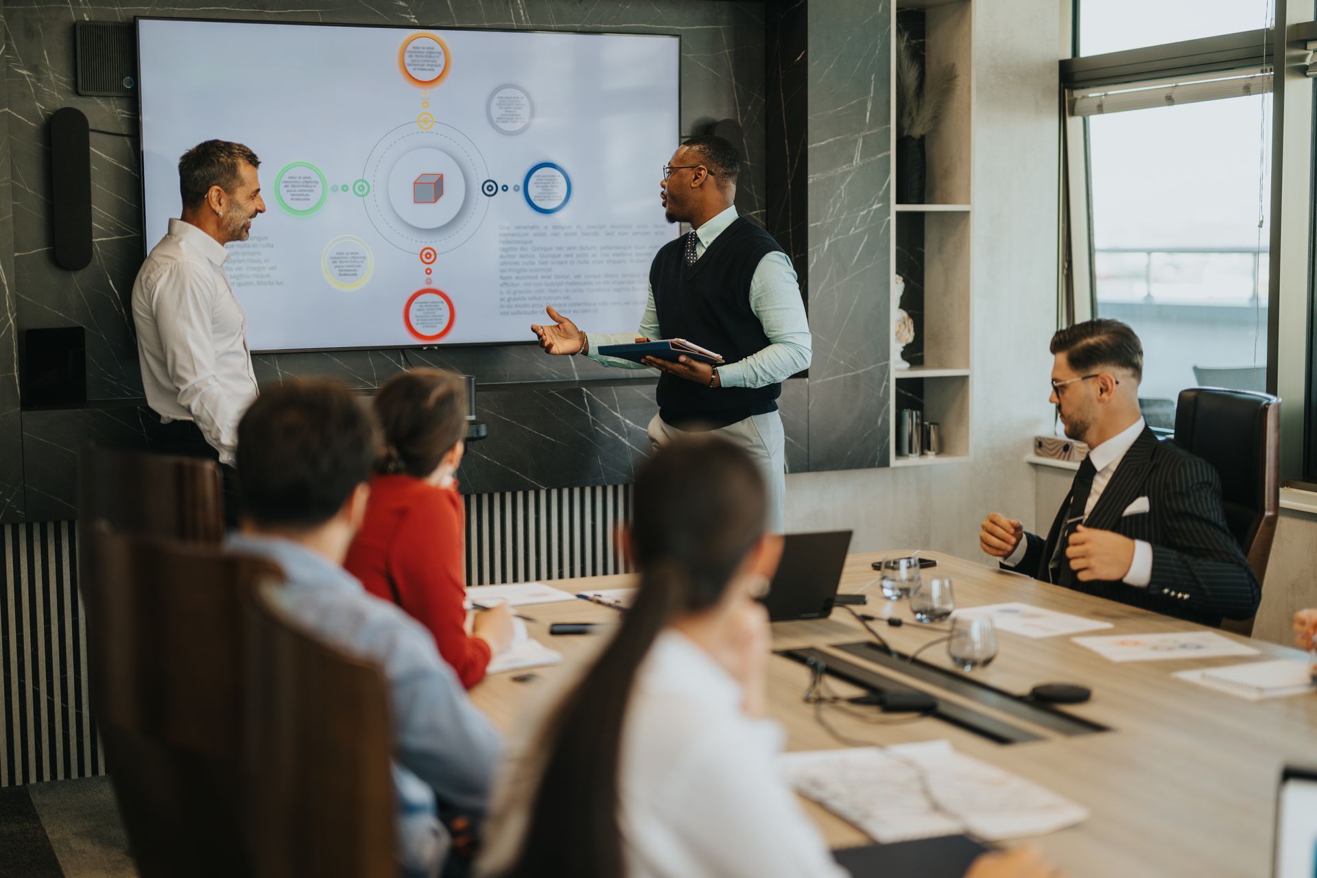 A business team meeting in an office conference room, with a speaker presenting a diagram on a large screen to colleagues.