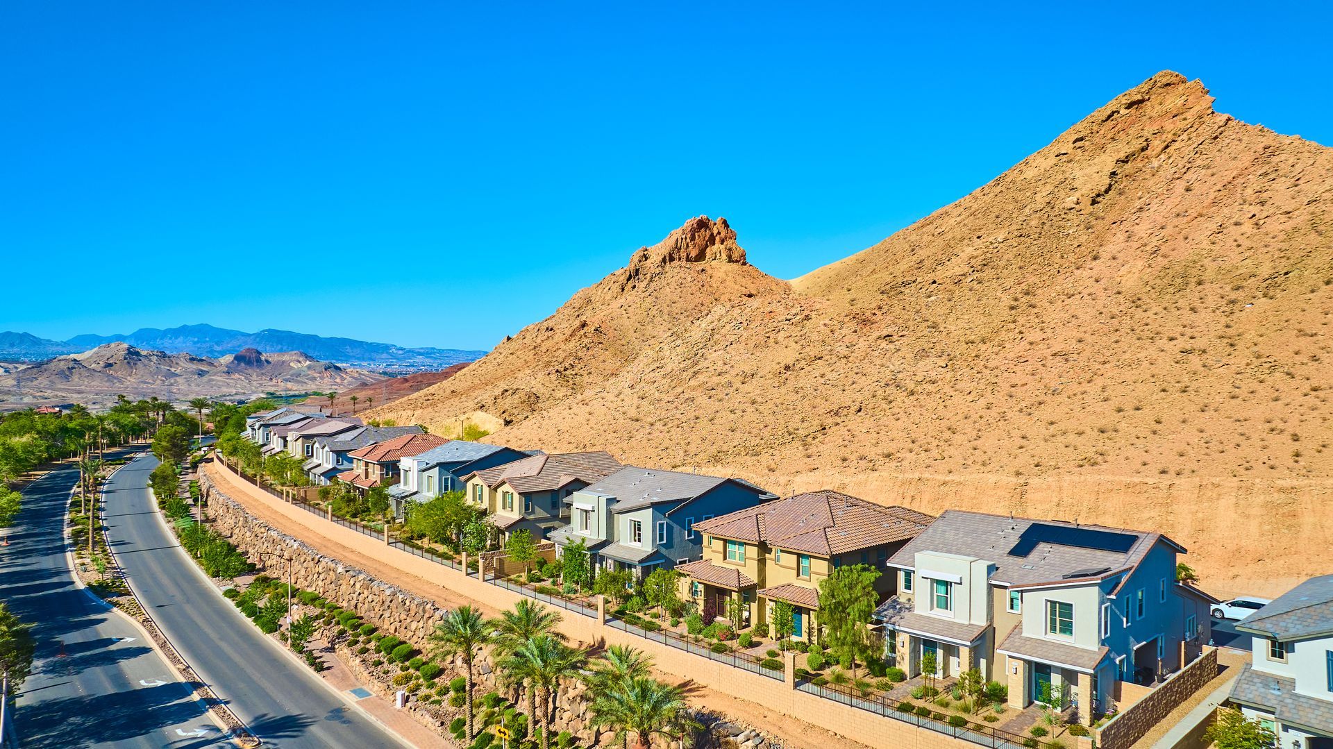 A row of suburban homes nestled against the base of a large, arid, tan-colored mountain under a clear blue sky.