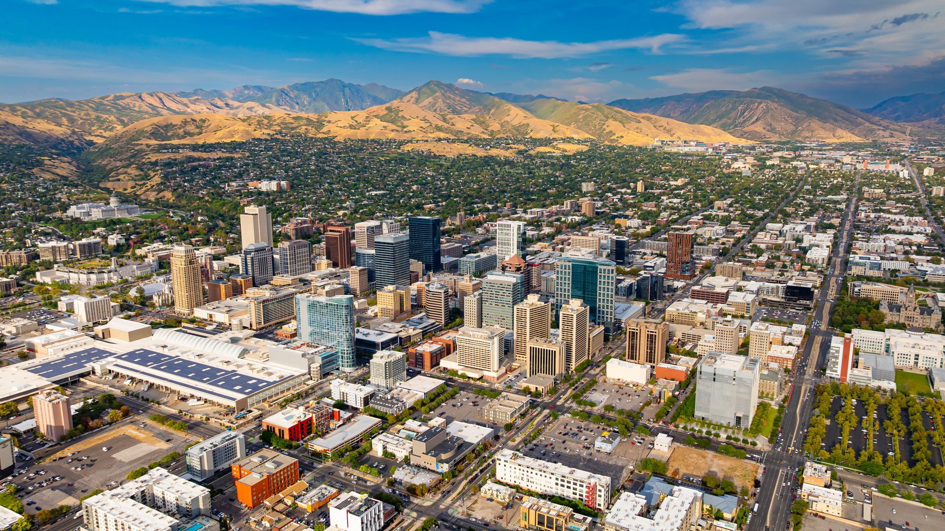 An aerial view of the Salt Lake City skyline and urban center, set against a backdrop of tan, sun-drenched mountains.