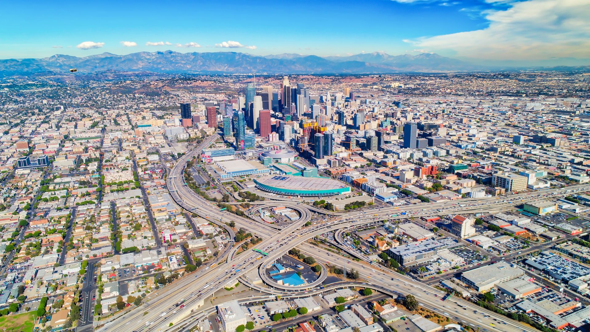 Aerial view of Los Angeles skyline and surrounding urban sprawl with a major freeway interchange under a blue sky.