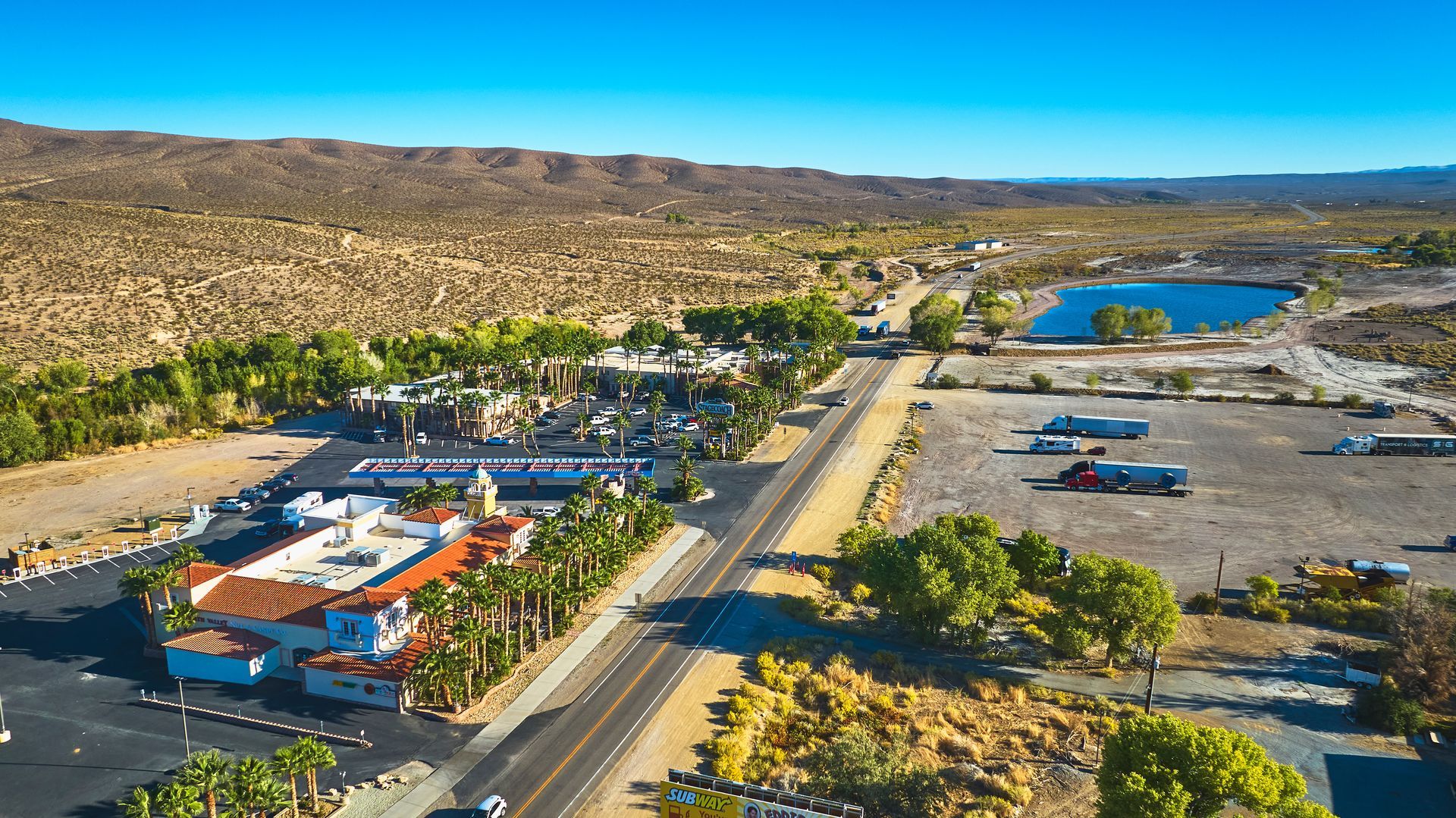 An aerial view of a desert town with a long, straight road stretching toward a mountain range under a clear blue sky.