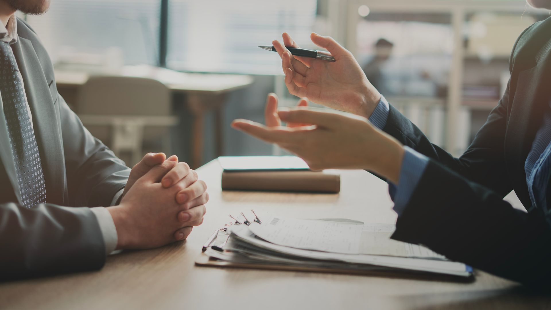 Two people in suits sitting across from each other at a desk with paperwork, engaged in a professional conversation.